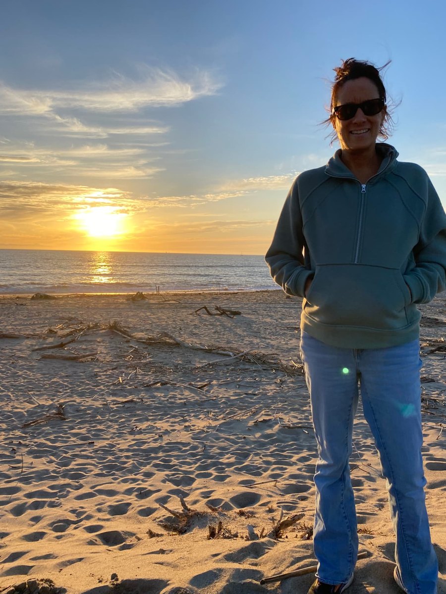 Cindy Costley standing on a sandy beach at sunset, wearing a sage-green half-zip pullover and sunglasses, with driftwood and the ocean behind her.